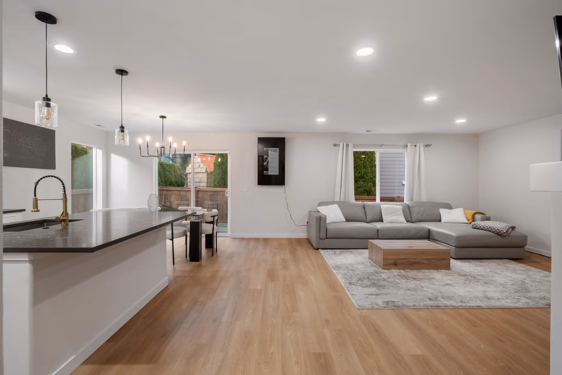 A modern open-concept living area featuring a kitchen with a dark countertop island and brass faucet on the left, a dining area with a table and chairs in the center near a sliding glass door, and a living room with a gray sectional sofa, a wooden coffee table on a light gray rug, and a window with white curtains on the right.