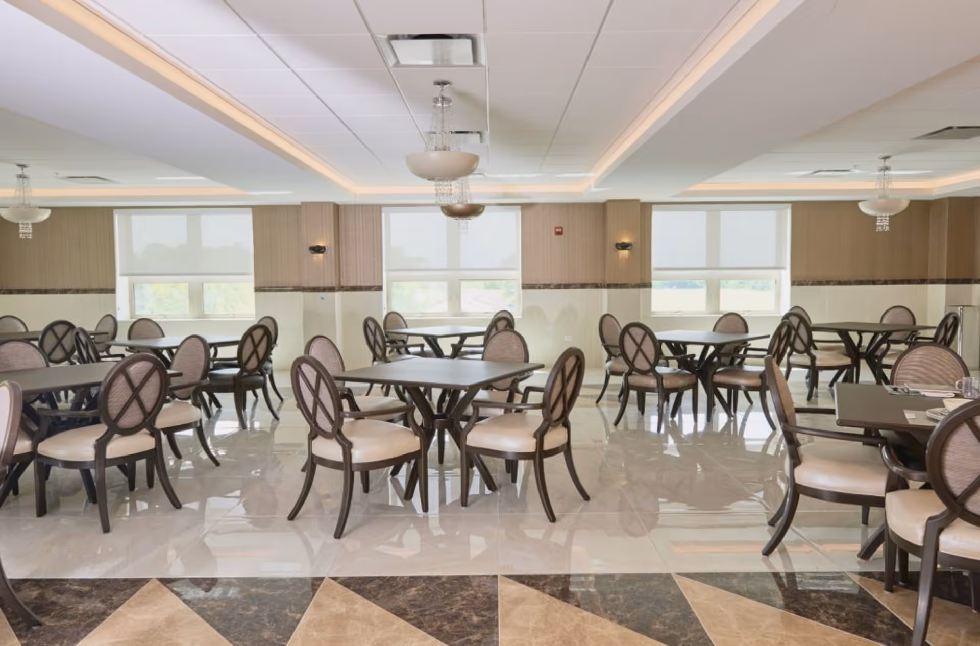A spacious dining room with multiple round tables and cushioned chairs arranged neatly. The room has large windows with white blinds, beige walls, and a polished tiled floor. Ceiling lights with decorative fixtures hang above the tables.