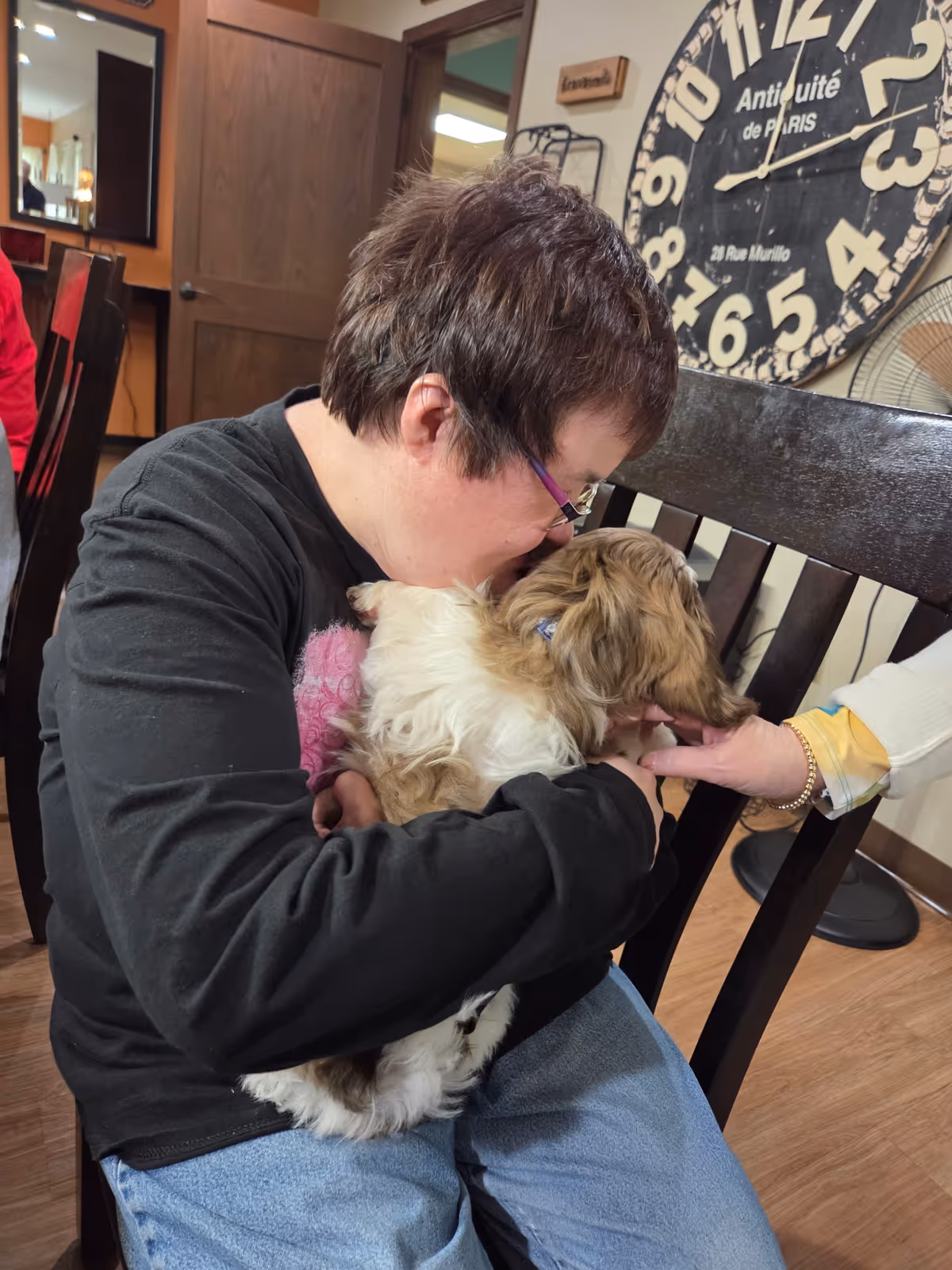 A person wearing glasses and a black long-sleeve shirt is sitting on a wooden chair indoors, holding and cuddling a small fluffy dog. Another person's hand is reaching out to pet the dog. In the background, there is a large decorative wall clock and a wooden door.