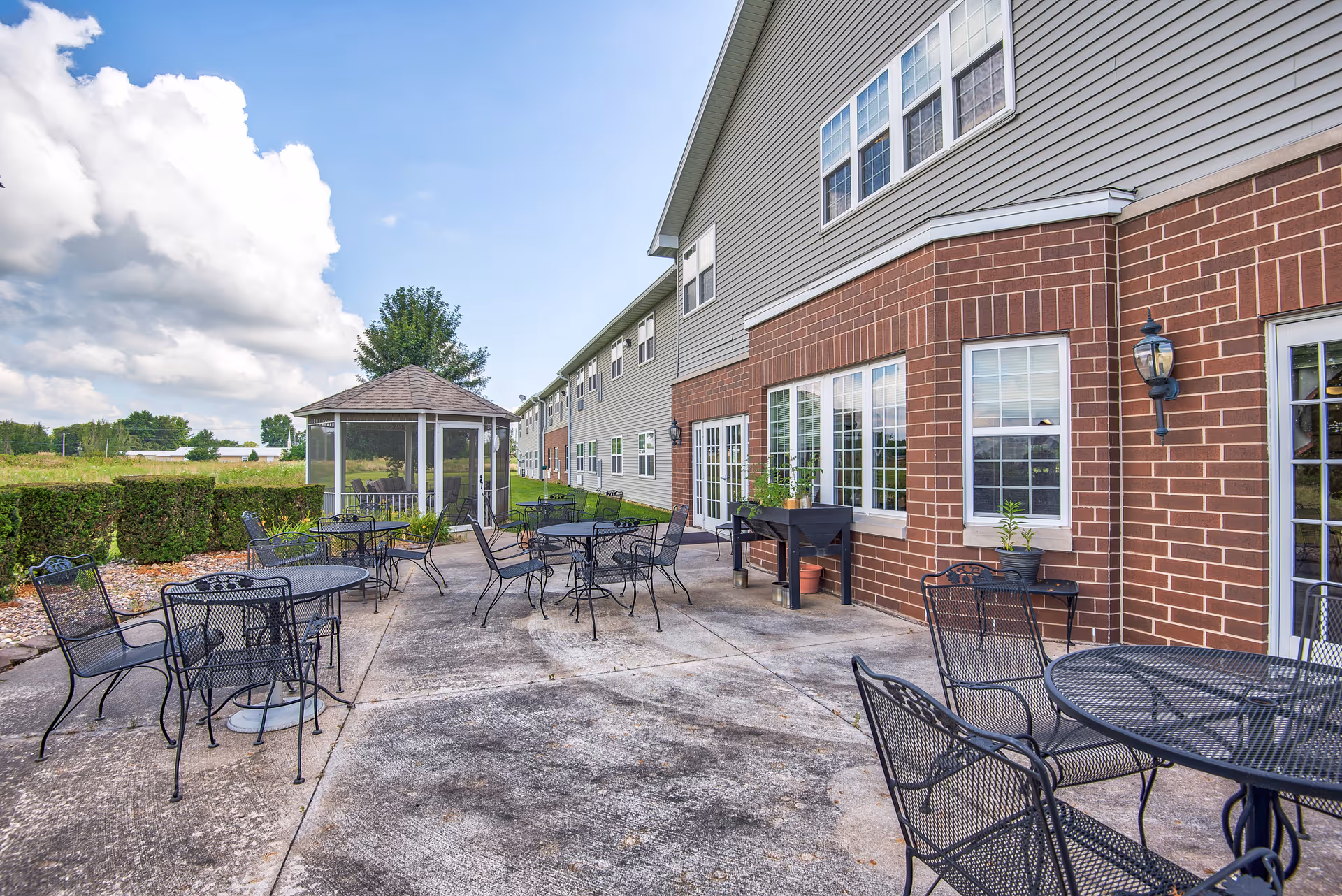 Outdoor patio area with multiple black metal tables and chairs on a concrete surface next to a brick and siding building. There is a small gazebo with mesh sides in the background, surrounded by greenery and a partly cloudy sky.