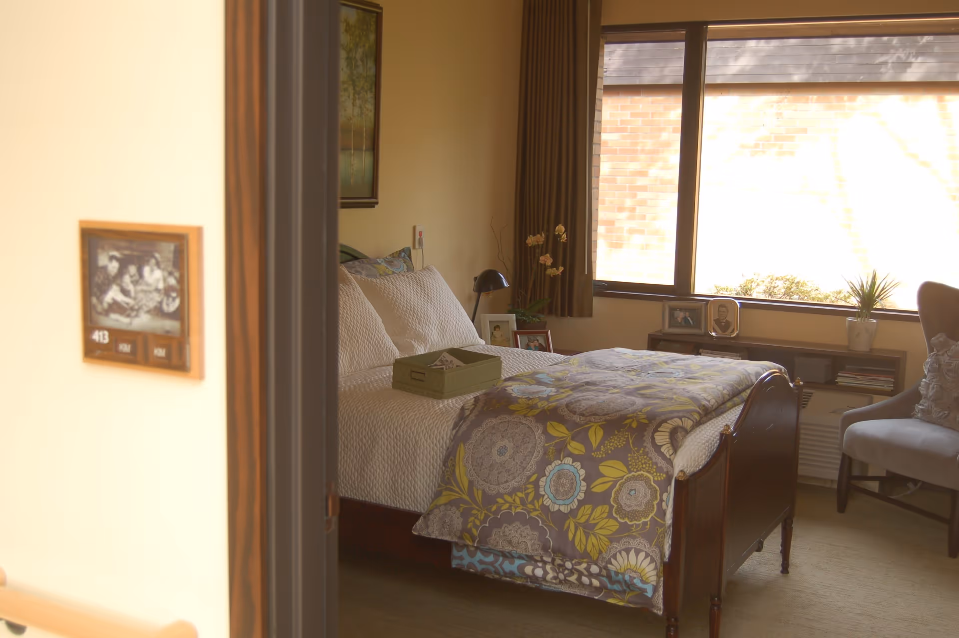 A cozy bedroom in a senior living facility with a neatly made bed featuring a floral patterned quilt and white pillows. The room has a large window with brown curtains, a small green basket on the bed, framed photos on a shelf under the window, a potted plant, and a comfortable armchair with a decorative pillow.