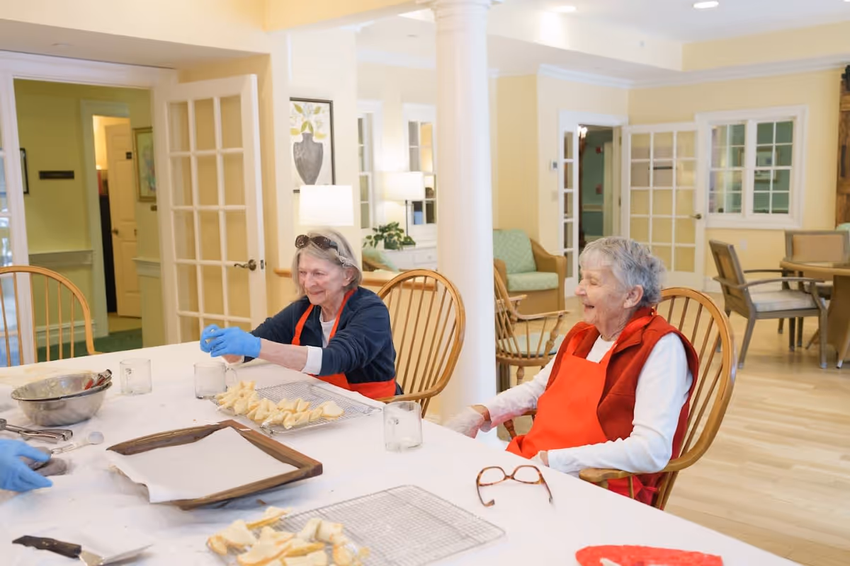 Two elderly women wearing red aprons sitting at a table in a bright room, preparing food with baking trays and dough pieces in front of them. The room has wooden chairs, light-colored walls, and glass-paned doors in the background.