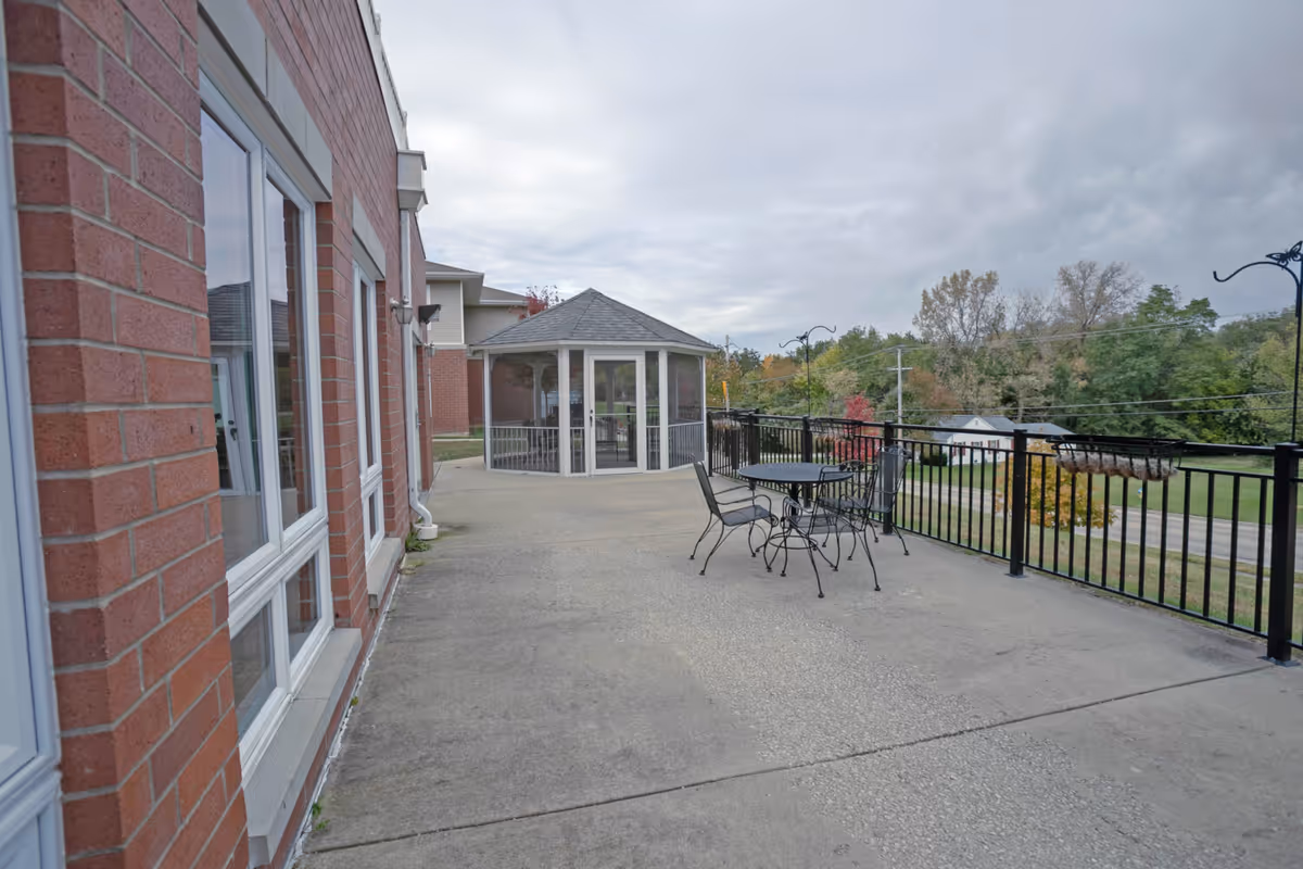 Outdoor patio area at Heritage Woods of Charleston featuring a concrete floor, black metal table with four matching chairs, black metal railing, and a small gazebo with screened windows. The patio overlooks a green landscape with trees and a road in the background under a cloudy sky.
