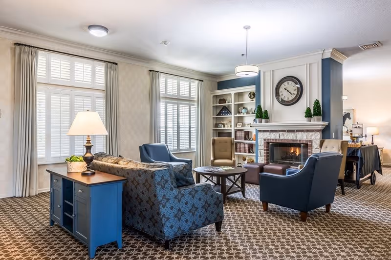 A cozy living room area in an assisted living facility featuring a patterned carpet, a blue patterned sofa, two blue armchairs, a beige armchair, a round wooden coffee table, and a fireplace with a clock above it. The room has large windows with white shutters and light curtains, a blue side table with a lamp and a plant, and built-in shelves with decorative items.
