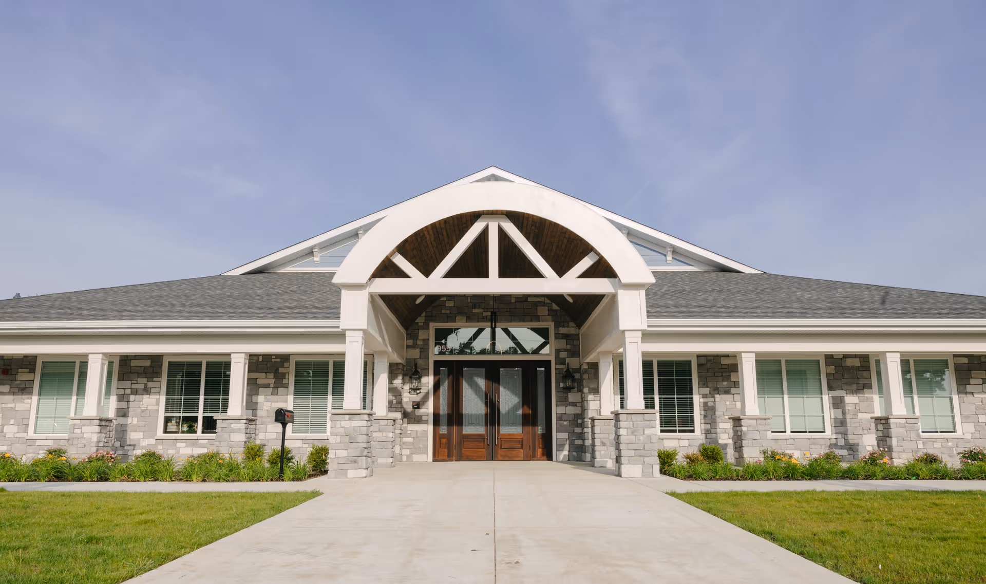 Front exterior view of a single-story building with a stone facade, large wooden double doors, and a covered entrance with white pillars and an arched roof structure. The building is surrounded by a well-maintained lawn and landscaping under a clear sky.