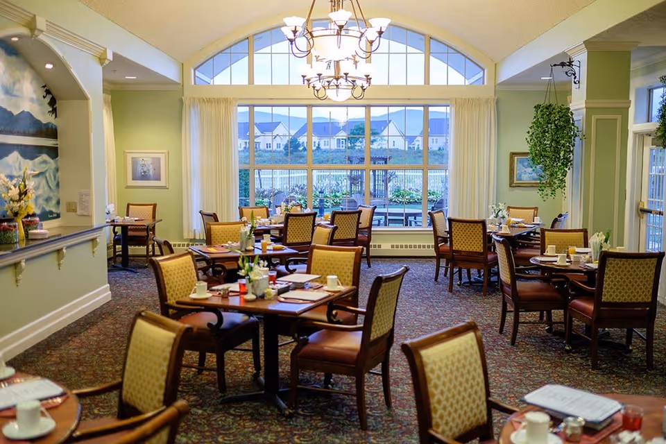 A bright dining room with multiple wooden tables and cushioned chairs arranged neatly. Each table is set with cups, glasses, and small flower arrangements. Large windows with white curtains allow natural light to fill the room, and a chandelier hangs from the ceiling. Outside the windows, houses and greenery are visible. The walls are painted light green, and there is a hanging plant on the right side.