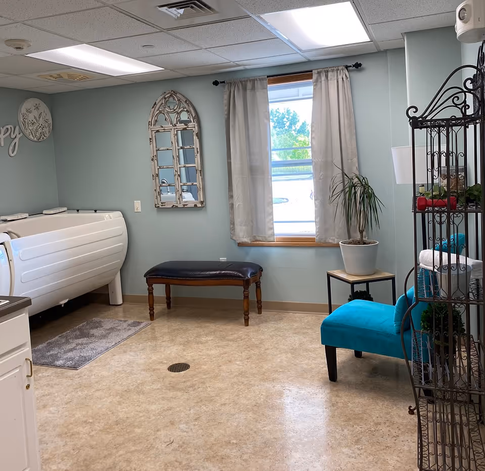A light blue room with a white walk-in bathtub on the left, a dark wooden bench with a black cushioned seat in the center, and a turquoise upholstered chair on the right. There is a window with beige curtains, a potted plant on a small table, a decorative mirror on the wall, and a metal shelving unit holding towels and plants.