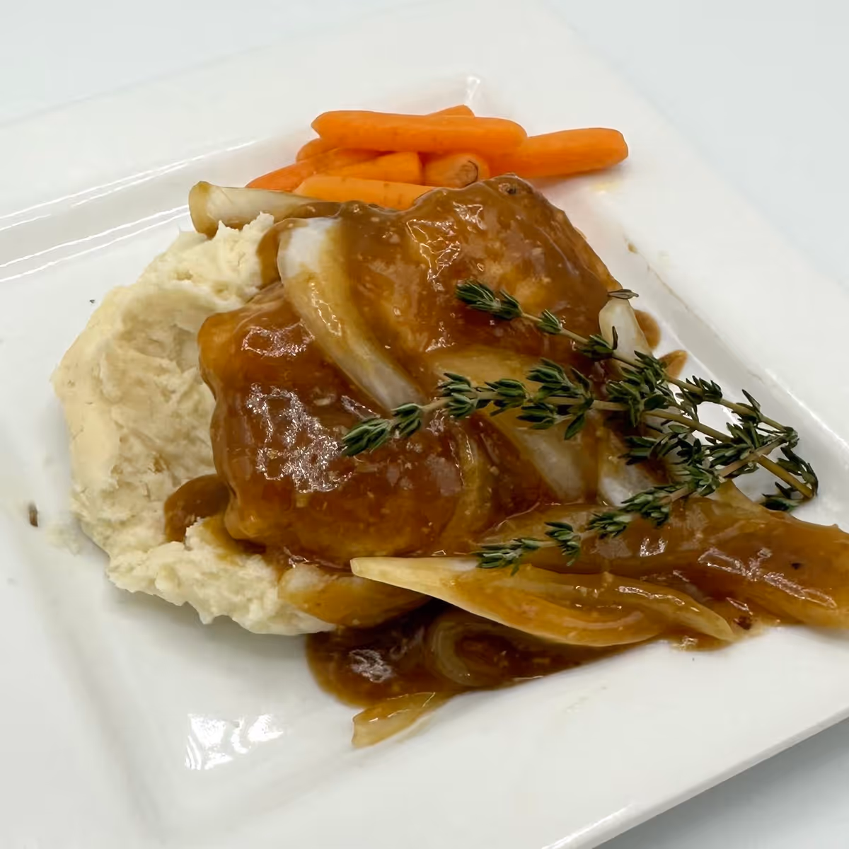 A plated meal consisting of mashed potatoes topped with brown gravy and onions, garnished with a sprig of thyme, served with cooked baby carrots on the side.