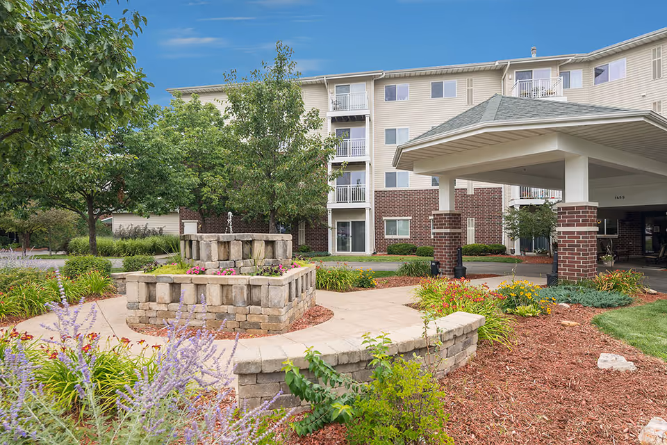 Outdoor view of a senior living facility with a multi-story building in the background, featuring balconies and a covered entrance supported by brick pillars. In the foreground, there is a stone fountain surrounded by landscaped flower beds and greenery under a clear blue sky.