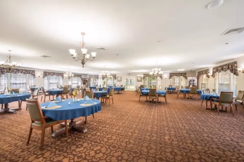 A spacious dining room with multiple round tables covered in blue tablecloths, each set with napkins and small flower vases. The room has patterned carpet flooring, several chandeliers hanging from the ceiling, and large windows with floral valances allowing natural light to fill the space.