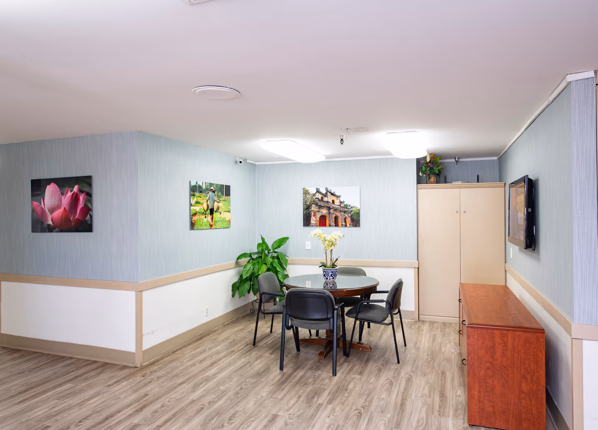 A small common area in a senior living facility with light blue walls and wood flooring. There is a round table with four black chairs around it, a potted plant on the table, and a larger green plant in the corner. The walls are decorated with three framed pictures, and there is a beige cabinet and a wooden sideboard along one wall. A flat-screen TV is mounted on the wall above the sideboard.
