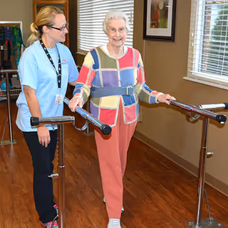 An elderly woman wearing a colorful patchwork sweater and pink pants is using parallel bars for walking assistance in a rehabilitation setting. A female caregiver in a light blue uniform is standing beside her, providing support and encouragement. The room has wooden flooring, large windows with blinds, and framed artwork on the wall.