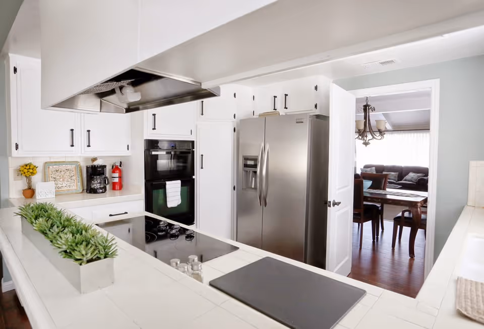 Bright kitchen with white cabinets, a stainless steel refrigerator, double oven, and an electric stovetop. A small rectangular planter with green plants is on the counter. Through an open door, a dining area with a wooden table, chairs, and a chandelier is visible, along with a living room with a sofa and large window.