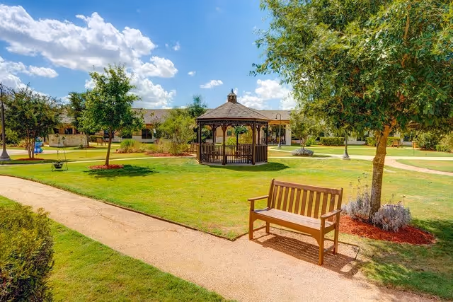 Outdoor garden area at The Auberge at Onion Creek featuring a wooden gazebo in the center, surrounded by green grass, trees, and walking paths. A wooden bench is placed near a tree on the right side of the image under a bright blue sky with scattered clouds.
