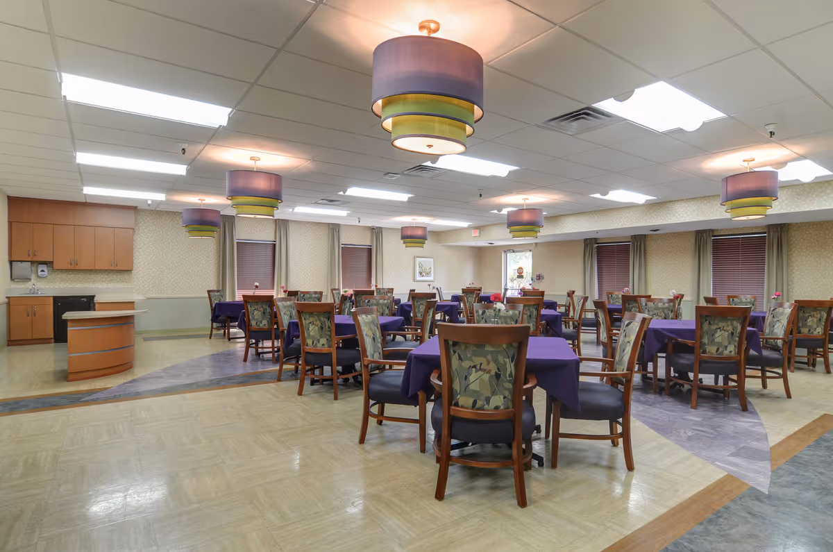 Spacious senior dining room with multiple tables draped in purple tablecloths, wooden chairs, and layered colorful pendant lights.