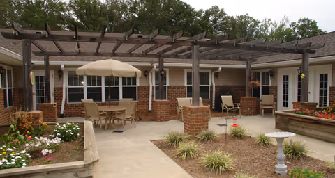 Outdoor patio area at Brookdale Monroe Square featuring a pergola with seating arrangements including a table with an umbrella and chairs, surrounded by flower beds and greenery.