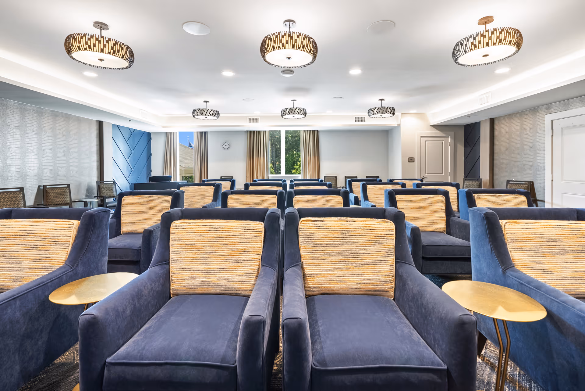 A bright, modern common area with multiple rows of blue armchairs featuring striped beige cushions, small round gold side tables, patterned ceiling lights, and large windows with beige curtains letting in natural light.