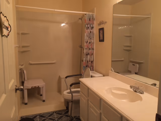 Bathroom with a walk-in shower featuring a white shower chair and grab bars. There is a toilet with arm supports next to a vanity with a sink and a large mirror above it. The shower curtain has a pattern, and the floor has a dark tile design.
