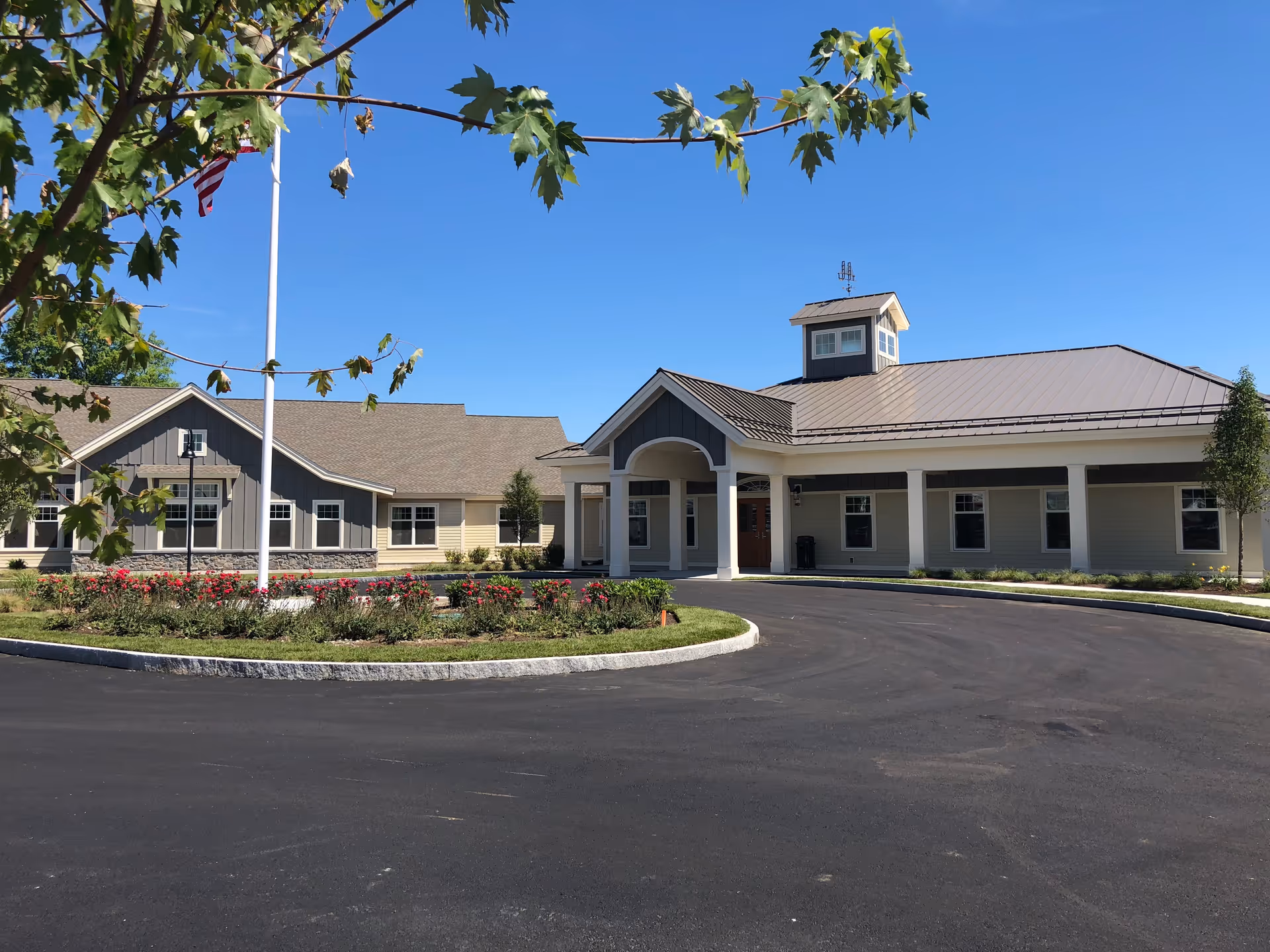 Front exterior of a single-story senior living facility with a covered entrance, circular driveway, landscaped beds, and an American flag.