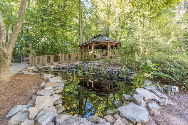 A serene gazebo surrounded by lush greenery and a small pond, with rocks lining the water's edge.