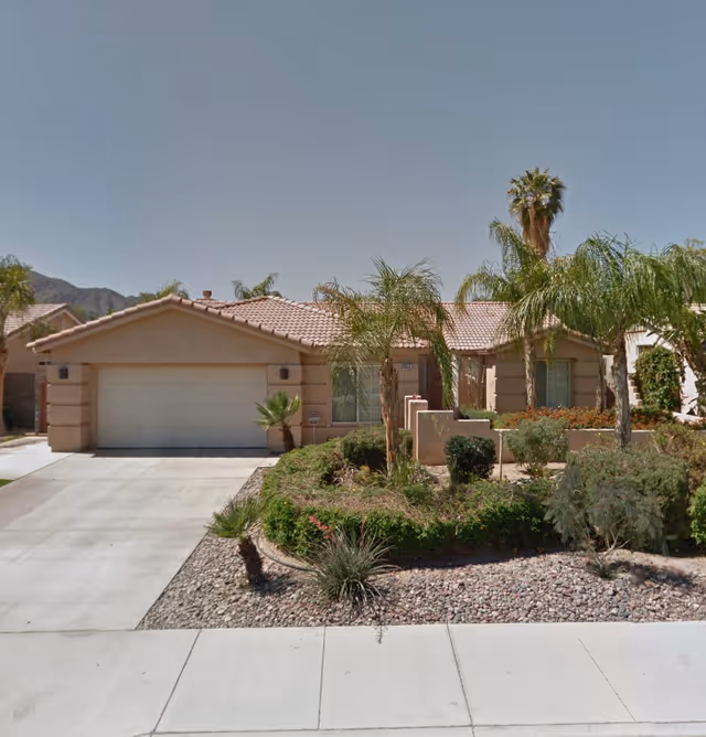 Single-story residential building with a tiled roof, a two-car garage, and a landscaped front yard featuring palm trees and various shrubs under a clear sky.