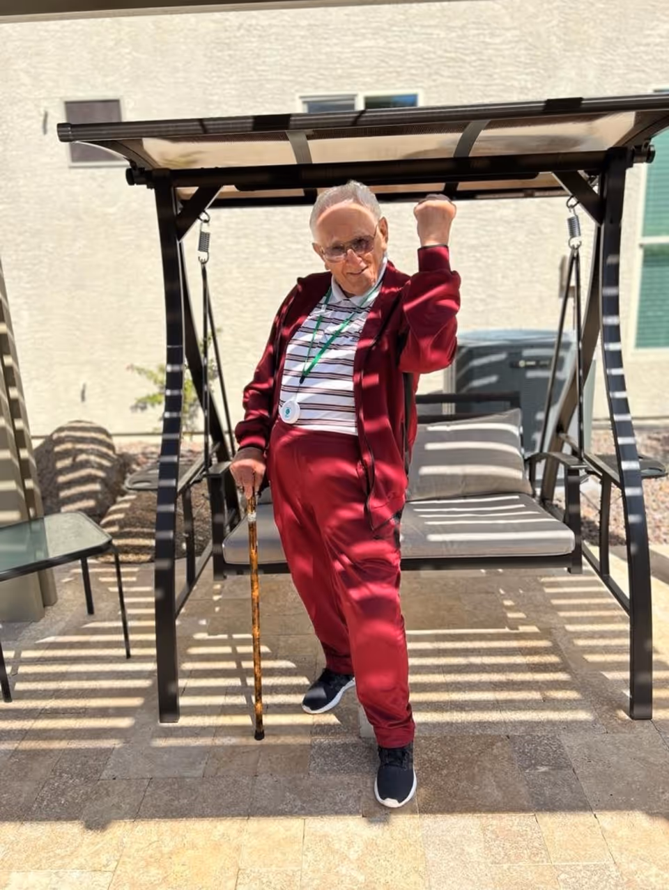 An elderly man wearing a red tracksuit and striped shirt stands outdoors on a tiled patio. He is holding a wooden cane in one hand and raising his other fist in a celebratory gesture. Behind him is a shaded swing bench with cushions, and the background shows a light-colored building wall with windows.