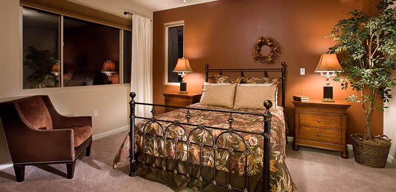 A cozy bedroom with a metal framed bed adorned with floral patterned bedding and pillows. There are two wooden nightstands on either side of the bed, each with a table lamp. A decorative wreath hangs on the wall above the bed. A brown upholstered chair is placed near a large window with white curtains. A potted plant is situated in the corner of the room, adding a touch of greenery.