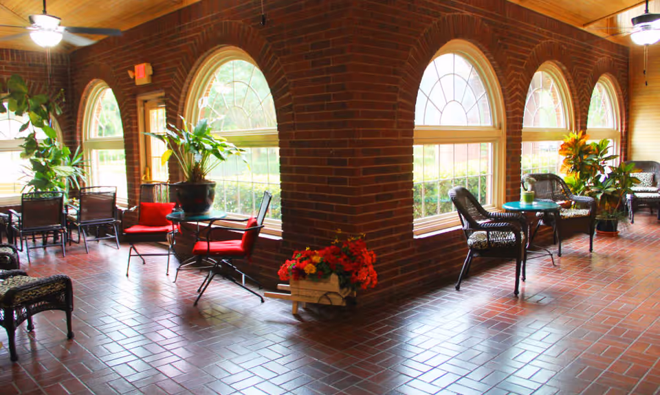 Sunlit brick-walled lounge with arched windows, wicker chairs, small tables, and potted plants.