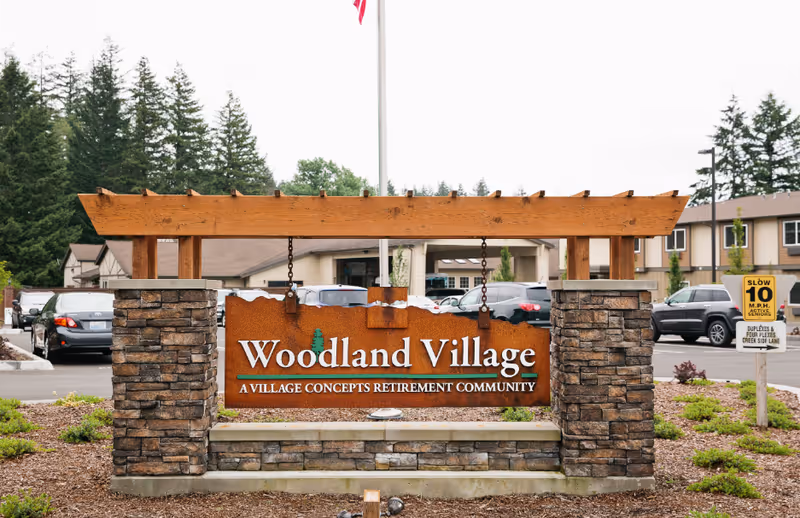 Entrance sign for Woodland Village, a Village Concepts retirement community, displayed on a stone and wood structure with parked cars and residential buildings in the background.