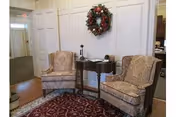 A cozy seating area with two patterned armchairs placed on a red patterned rug. Between the chairs is a small wooden table with a decorative item on it. A festive wreath hangs on the white paneled wall behind the chairs. The space appears to be part of an interior common area with a hallway visible in the background.