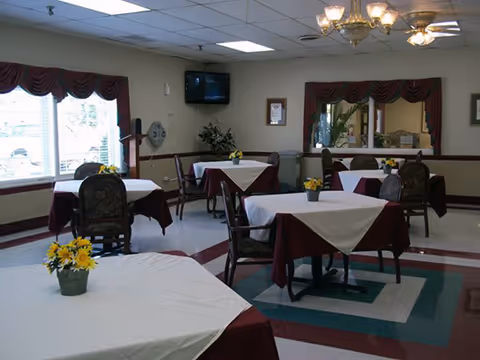 A dining room with several tables covered with white and burgundy tablecloths, each table decorated with a small pot of yellow flowers. The room has large windows with burgundy valances, a wall-mounted TV, a ceiling fan, and a chandelier. There is a large mirror on one wall reflecting part of the room.