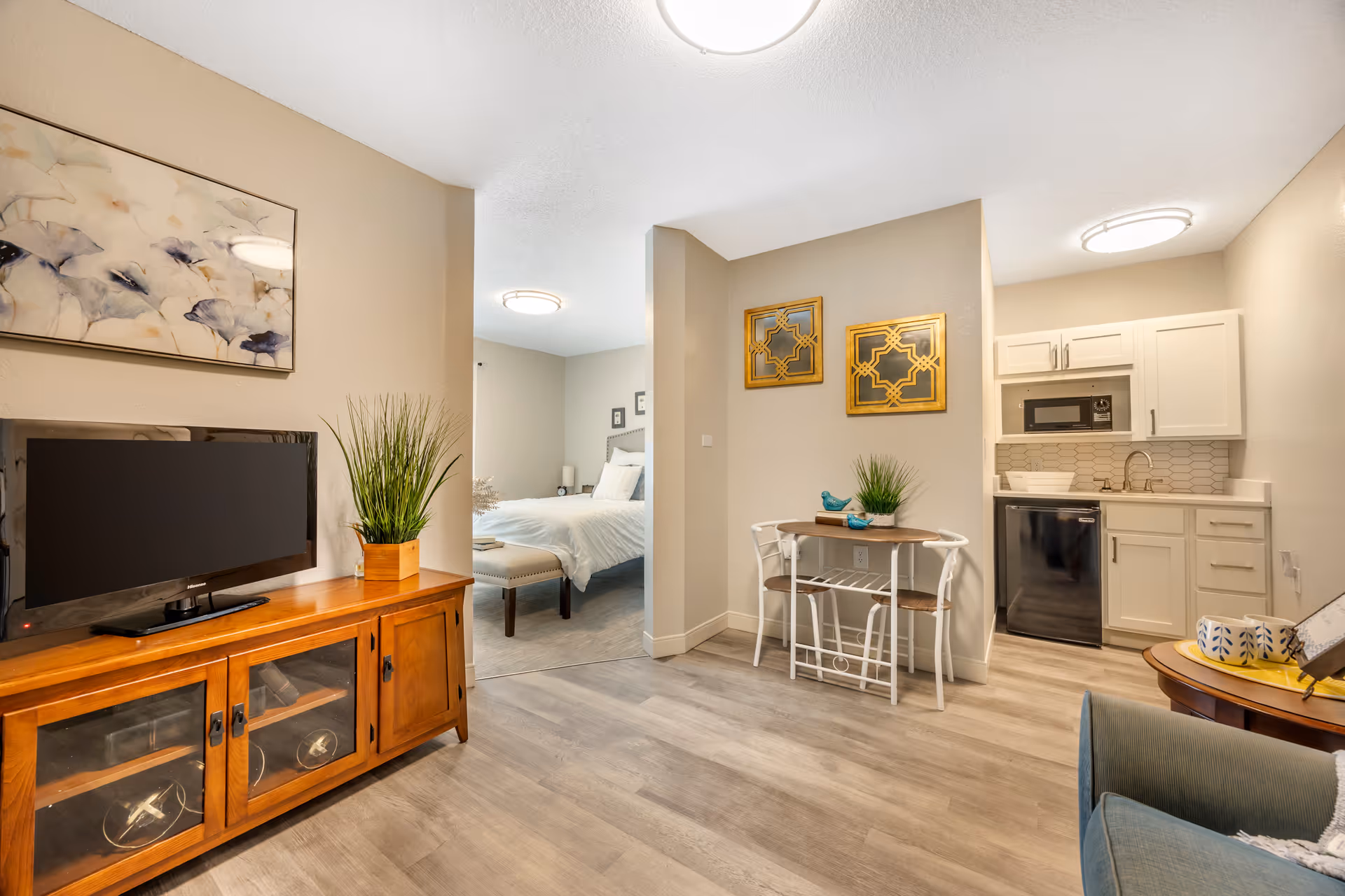 Interior view of a senior living facility apartment showing a small living area with a wooden TV stand and flat screen TV, a small dining table with two chairs, a kitchenette with white cabinets, a microwave, and a mini fridge. In the background, a bedroom with a bed and nightstand is visible. The walls are beige, and the floor is light wood.