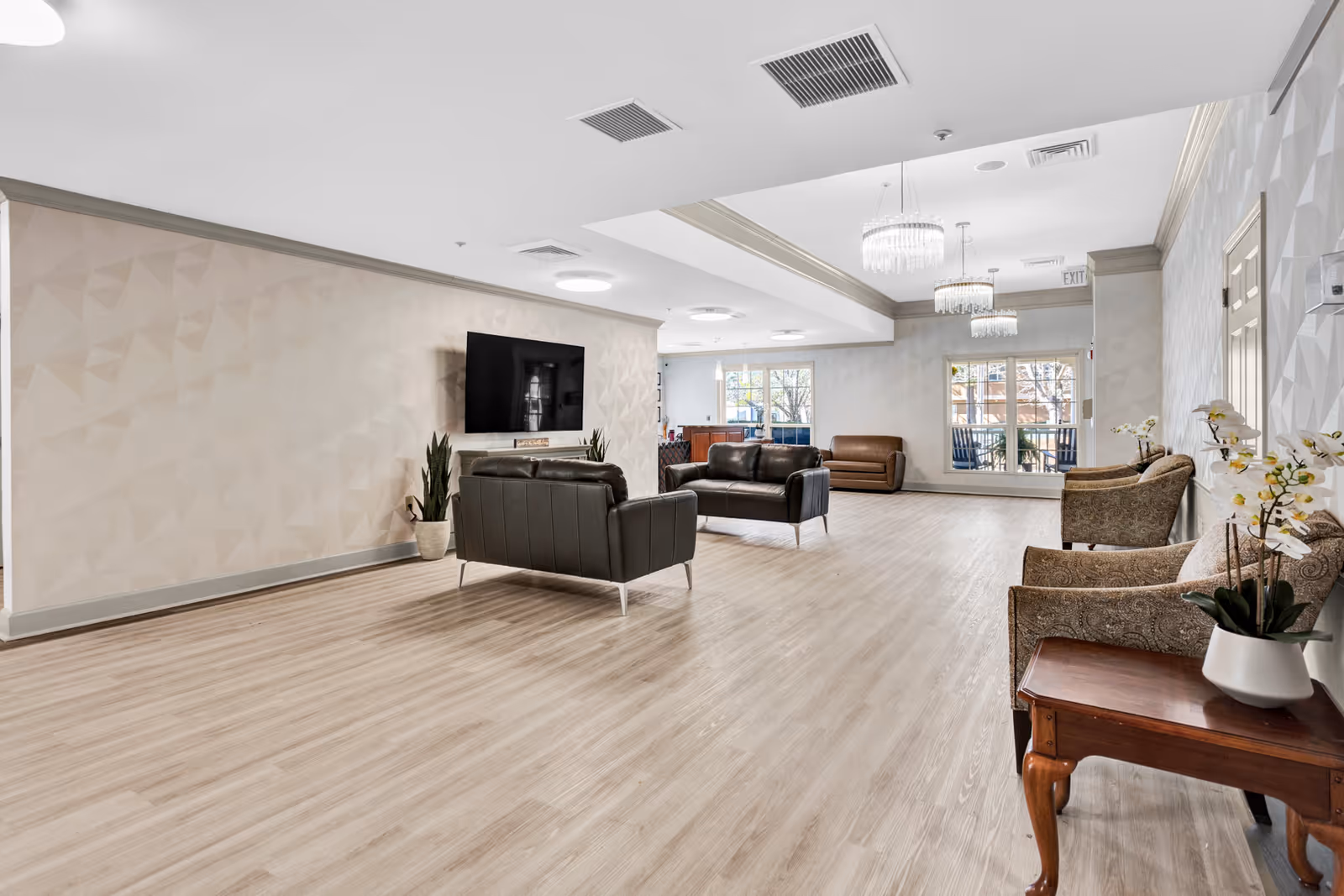 A spacious and well-lit living room area in an assisted living facility with light wood flooring, beige textured walls, and a mounted flat-screen TV. The room features two black leather sofas facing the TV, a brown leather chair near large windows, and three patterned armchairs along the right wall next to a wooden side table with a white vase and flowers. Modern chandeliers hang from the ceiling, and there are potted plants near the TV.
