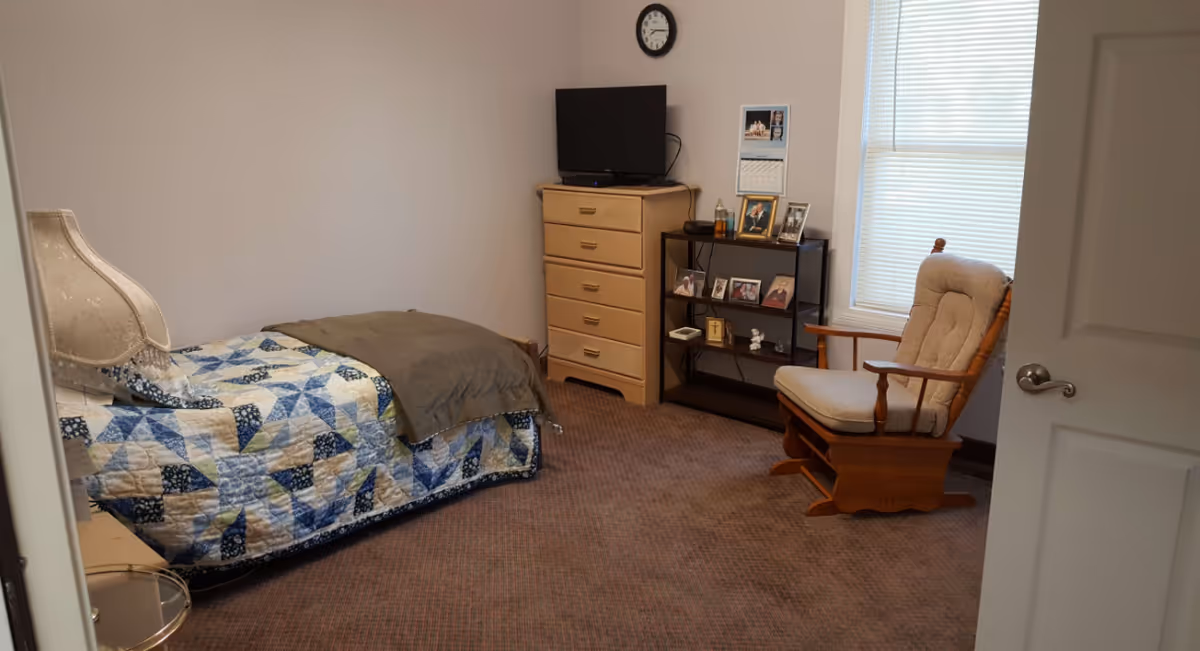 A small bedroom with a single bed covered with a blue and white patchwork quilt and a gray blanket. There is a wooden rocking chair with a cushioned seat near a window with blinds. A wooden dresser with a TV on top and a black shelving unit with framed photos and decorative items are against the wall. A clock is mounted above the TV on the wall.