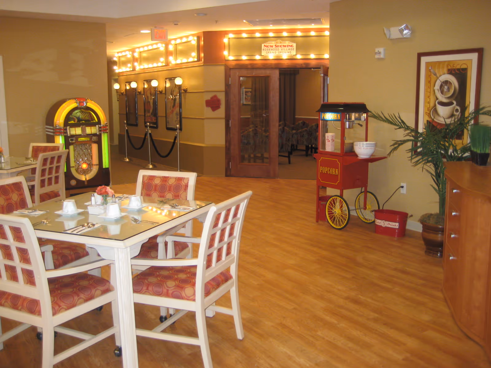 A dining area with a table set for four, featuring white chairs with red patterned cushions. In the background, there is a vintage jukebox, a popcorn machine on wheels, and a hallway with illuminated marquee lights and framed posters. The floor is wooden, and there are plants and decorative artwork on the walls.