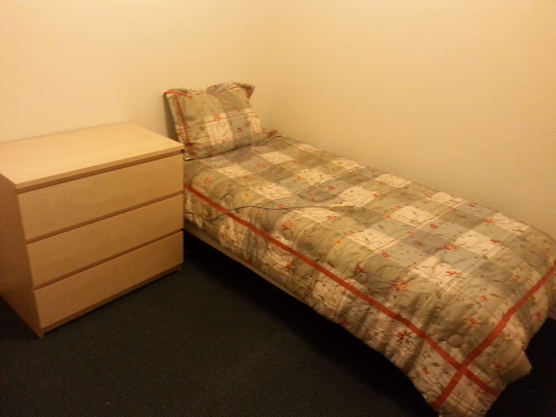 A small bedroom with a single bed covered in a floral patterned quilt and matching pillow. Next to the bed is a light wooden three-drawer dresser. The walls are plain and light-colored, and the floor is carpeted in a dark color.
