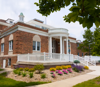 Exterior view of a brick building with white columns and a white railing around a raised porch area. The building has multiple windows and a small cupola on the roof. There are landscaped flower beds with colorful flowers and green shrubs in front, and trees partially frame the image.