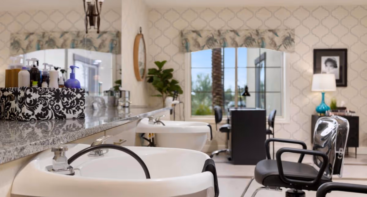 Interior of a salon area with two white wash basins in the foreground, a countertop with various hair care products, and salon chairs. In the background, there is a window with a view of palm trees and a desk with chairs. The room has patterned wallpaper and a lamp on a side table.