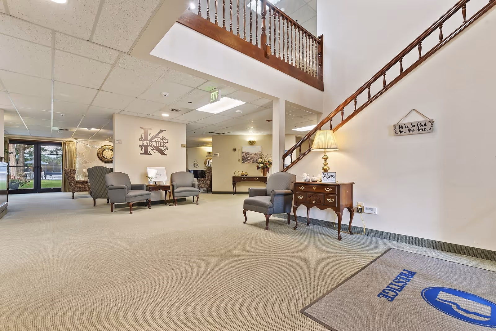 A spacious and well-lit senior living facility common area with beige carpeting, several gray upholstered armchairs, a wooden side table with a lamp, and a staircase with wooden railing. The wall features a decorative sign that says 'We're So Glad You Are Here.' Large windows and glass doors provide a view of the outdoor area with trees and seating. The wall also displays a large decorative letter 'K' with the word 'Karcher' underneath.