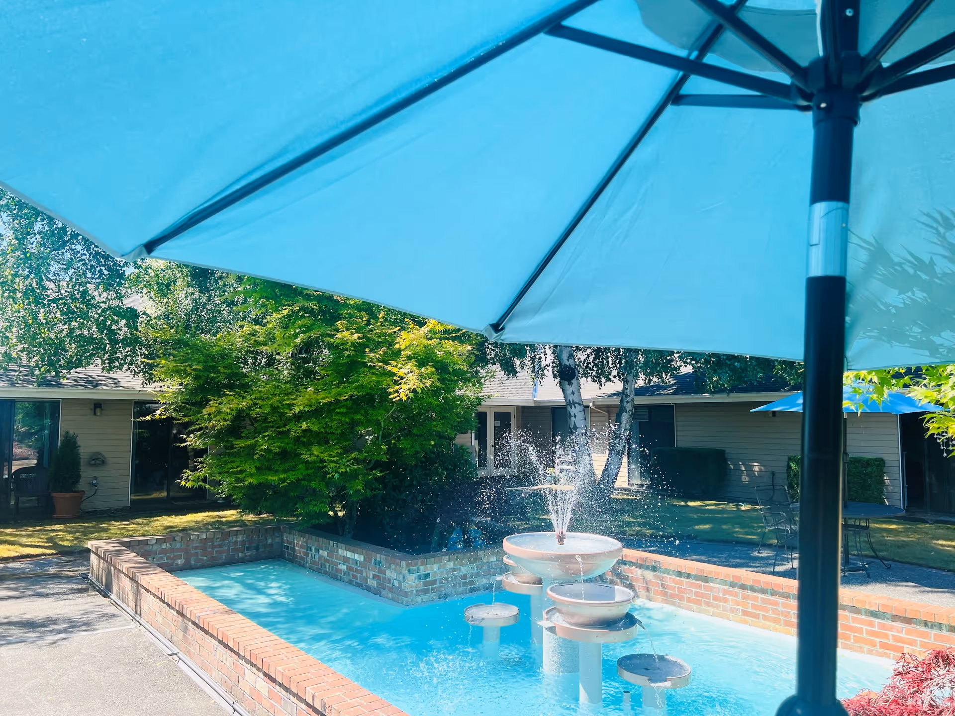 Outdoor courtyard area at Cap Sante Court Retirement Community featuring a brick-lined water fountain with multiple tiers spraying water, surrounded by greenery and trees. A large blue patio umbrella is partially visible in the foreground, shading part of the seating area.