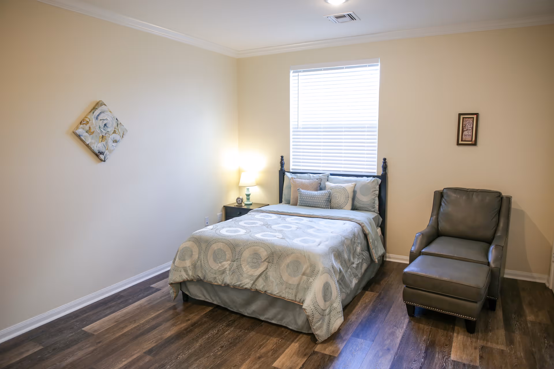 A cozy bedroom with a bed covered in a light blue patterned comforter and multiple pillows. There is a nightstand with a lamp and clock next to the bed. A gray armchair with an ottoman is positioned to the right of the bed. The room has wooden flooring, beige walls, a window with white blinds, and two pieces of wall art.