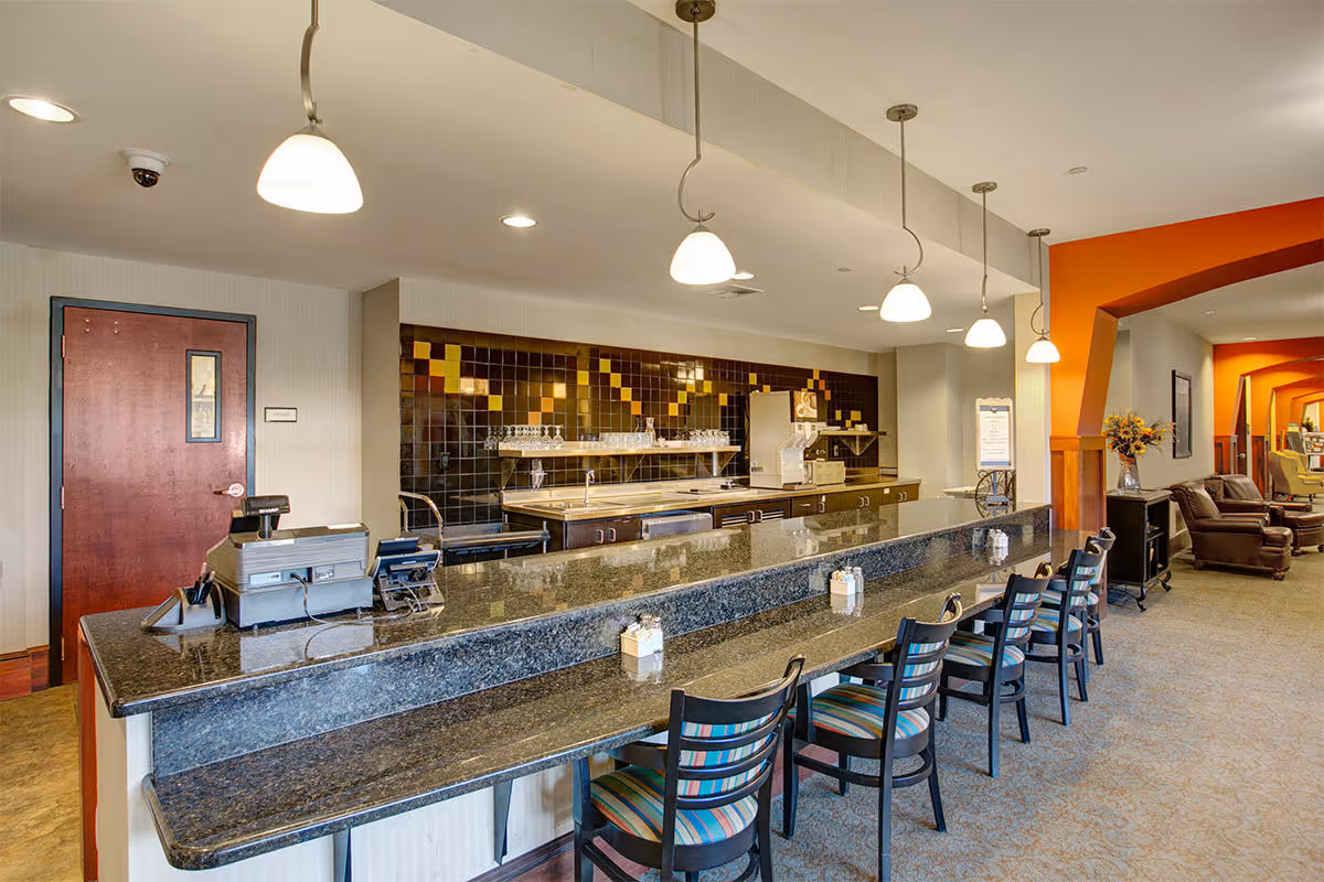 Interior view of a senior living facility's dining or common area featuring a long granite countertop with several chairs lined up. Behind the counter is a kitchen area with shelves holding glasses and a beverage dispenser. The space is well-lit with hanging pendant lights and has a warm color scheme with orange accents and comfortable seating visible in the background.