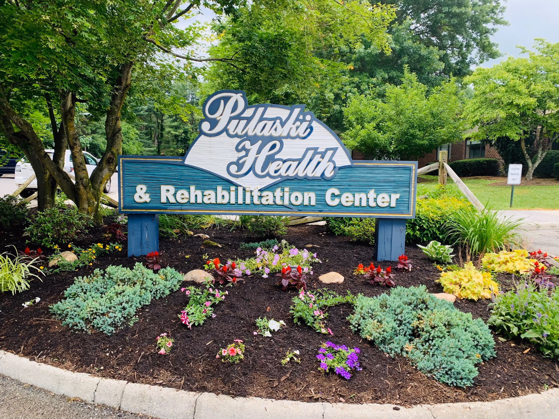 A landscaped garden area with various green shrubs and colorful flowers surrounding a large wooden sign that reads 'Pulaski Health & Rehabilitation Center'. Trees and a parking lot are visible in the background.
