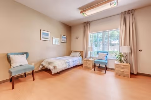 A bright and clean bedroom with a single bed against the wall, two blue upholstered chairs, two wooden nightstands with lamps, a large window with beige curtains, and three framed pictures on the wall.