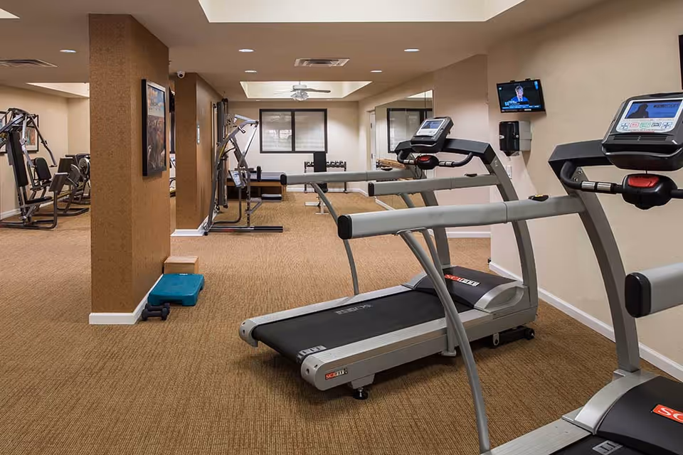 Interior view of a fitness room in a senior living facility with two treadmills in the foreground, various exercise machines in the background, a wall-mounted TV, and a large mirror on the right wall.