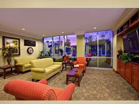 Comfortable living room/lounge with sofas and armchairs facing a TV and large windows overlooking a courtyard at dusk.