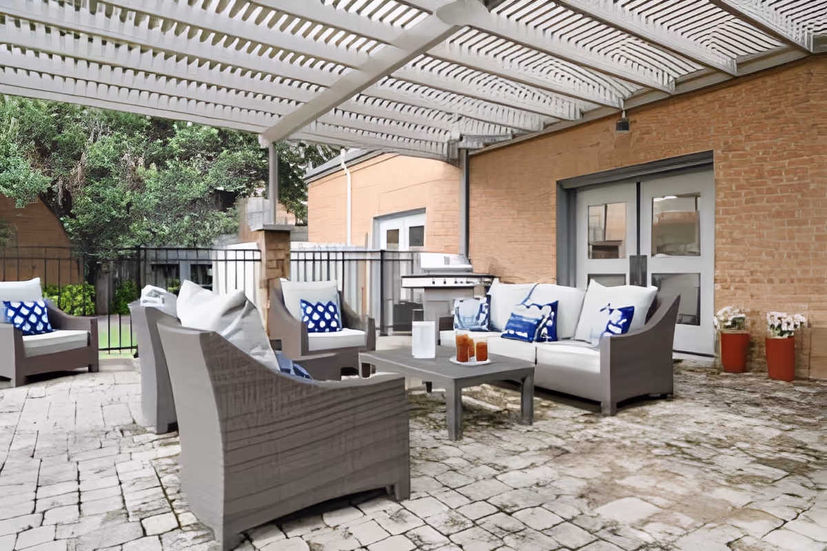 Outdoor patio area with cushioned seating including a sofa and armchairs arranged around a coffee table with drinks. The patio has a white pergola overhead, stone flooring, and a brick wall with double doors. There are potted plants and greenery visible in the background.