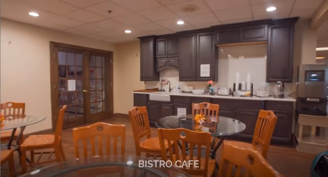 Small bistro-style dining area with round glass-top tables, wooden chairs, and a kitchenette in the background.