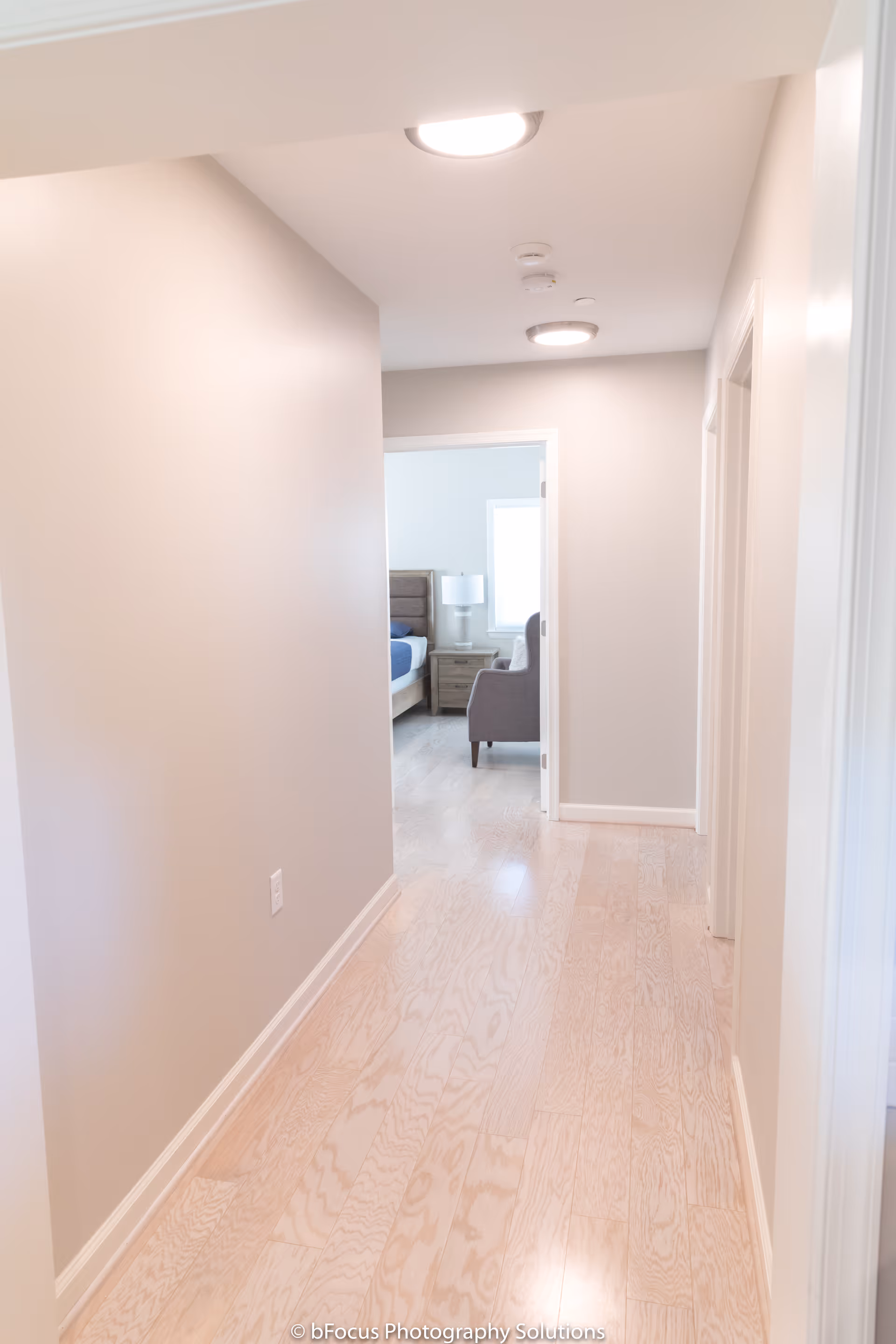 A bright hallway with light wooden flooring and beige walls leading to a bedroom. The bedroom has a bed with a blue blanket, a nightstand with a lamp, and a gray armchair near a window.