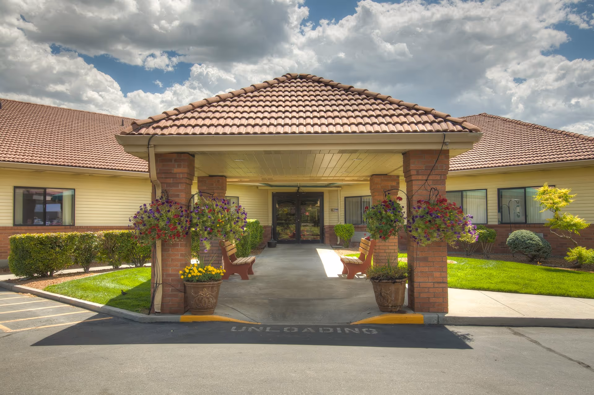 Entrance of Bridgeview Estates senior living facility with a covered driveway supported by brick pillars, hanging flower baskets, benches, and well-maintained landscaping under a partly cloudy sky.